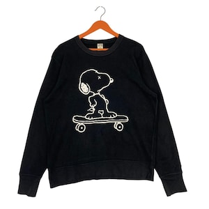 May include: Black sweatshirt featuring a white graphic of Snoopy skateboarding. The long-sleeved crew neck top is hanging on a wooden hanger. The UT logo is visible on the neckline.