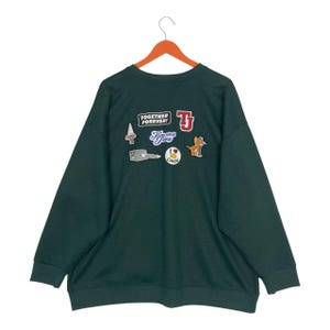 May include: Dark green sweatshirt with multiple Tom and Jerry cartoon patches. Patches include the words "TOGETHER FOREVER!" and "Tom and Jerry", along with cartoon images of the characters and cheese. The sweatshirt is hanging on a wooden hanger.