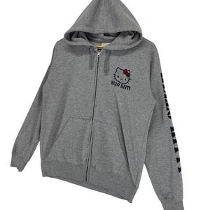 May include: A gray zip-up hoodie featuring a Hello Kitty graphic with a red bow and the words "Hello Kitty" in black. The left sleeve has black text. The hoodie has a hood and a front pocket.