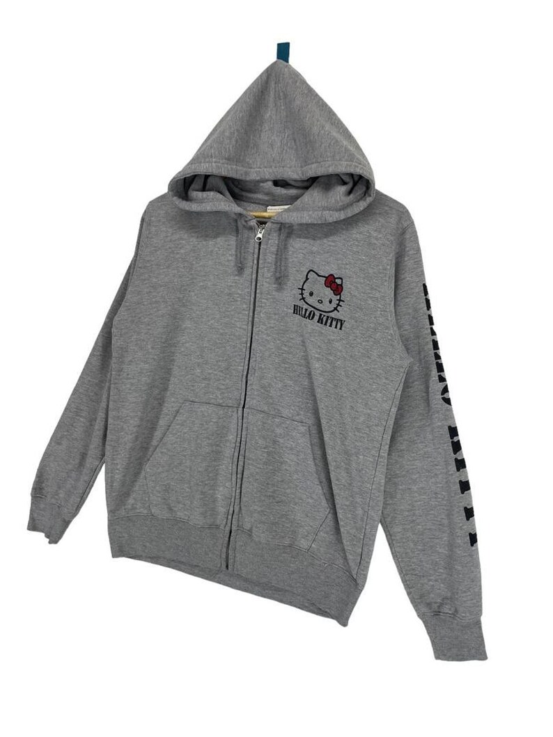 May include: A gray zip-up hoodie featuring a Hello Kitty graphic with a red bow and the words "Hello Kitty" in black. The left sleeve has black text. The hoodie has a hood and a front pocket.