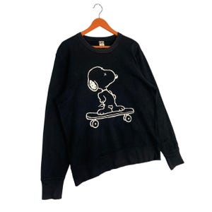 May include: Black crew neck sweatshirt featuring a white graphic of Snoopy skateboarding. The long-sleeved top is hanging on a wooden hanger. The sweatshirt has a ribbed neckline and cuffs.