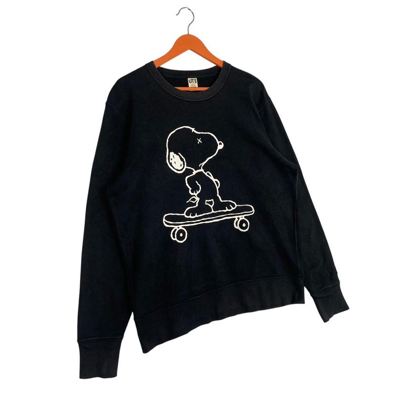 May include: Black crew neck sweatshirt featuring a white graphic of Snoopy skateboarding. The long-sleeved top is hanging on a wooden hanger. The sweatshirt has a ribbed neckline and cuffs.