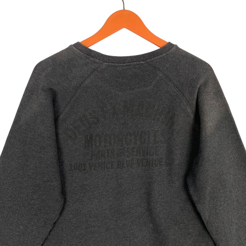 Pode incluir: Camisola cinza escuro com gola redonda e mangas raglan. A parte de tr&aacute;s da camisola apresenta uma impress&atilde;o preta desbotada que diz "Deus Ex Machina Motorcycles Parts & Service 1001 Venice Blvd Venice."