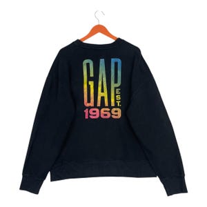 May include: Black crew neck sweatshirt with a rainbow gradient "GAP" logo on the back. The text "EST. 1969" is below the logo. The sweatshirt is suspended from an orange hanger.