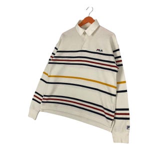 May include: A long-sleeve off-white polo shirt featuring horizontal stripes in navy blue, dark red, and mustard yellow. The shirt has a collar, a button placket, and the FILA logo embroidered in navy blue on the left chest and on the left cuff.