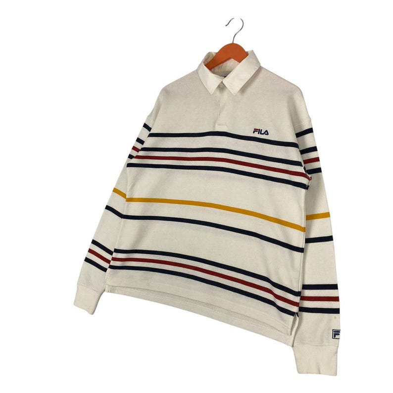 May include: A long-sleeve off-white polo shirt featuring horizontal stripes in navy blue, dark red, and mustard yellow. The shirt has a collar, a button placket, and the FILA logo embroidered in navy blue on the left chest and on the left cuff.