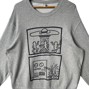 May include: Gray crewneck sweatshirt featuring a black graphic of a UFO beaming figures, and a control panel with a figure. The sweatshirt is hanging on a wooden hanger.