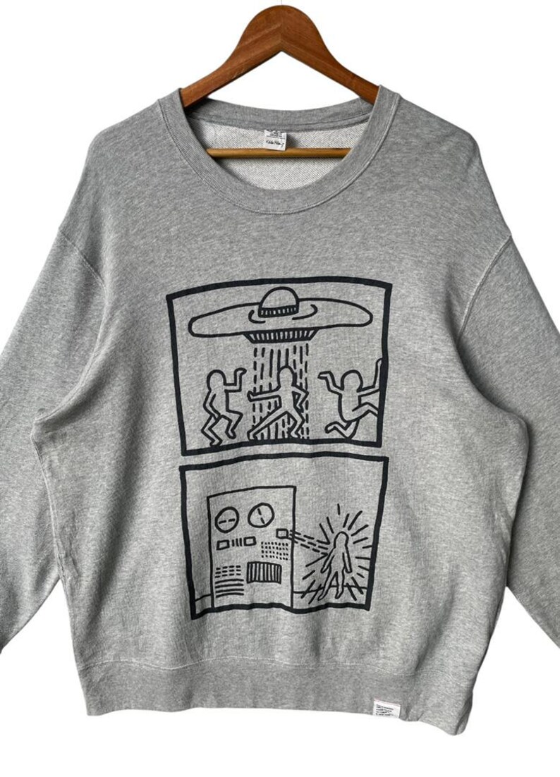 May include: Gray crewneck sweatshirt featuring a black graphic of a UFO beaming figures, and a control panel with a figure. The sweatshirt is hanging on a wooden hanger.