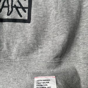 May include: Close-up of a gray sweatshirt featuring a black graphic design in a square frame. The bottom of the sweatshirt has a tag with the text "KEITH HARING: FIRST MAJOR EXHIBITION, OCTOBER 1982 IN NEW YORK CITY."
