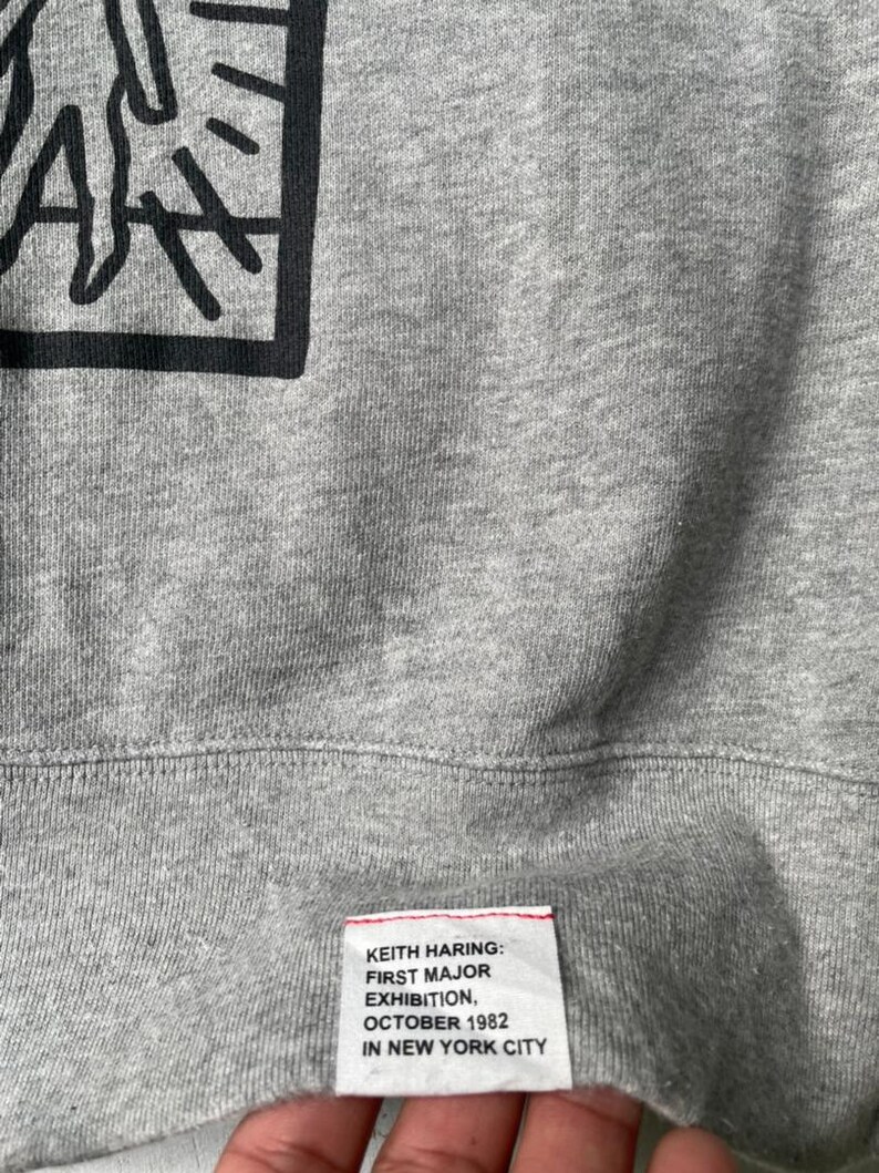 May include: Close-up of a gray sweatshirt featuring a black graphic design in a square frame. The bottom of the sweatshirt has a tag with the text "KEITH HARING: FIRST MAJOR EXHIBITION, OCTOBER 1982 IN NEW YORK CITY."