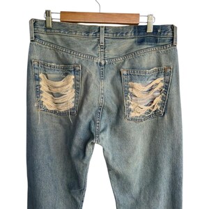 May include: Light blue denim jeans with distressed detailing on the back pockets. The jeans are hanging on a wooden hanger. The pockets have a shredded design, revealing the fabric underneath. The jeans are a classic, casual style.
