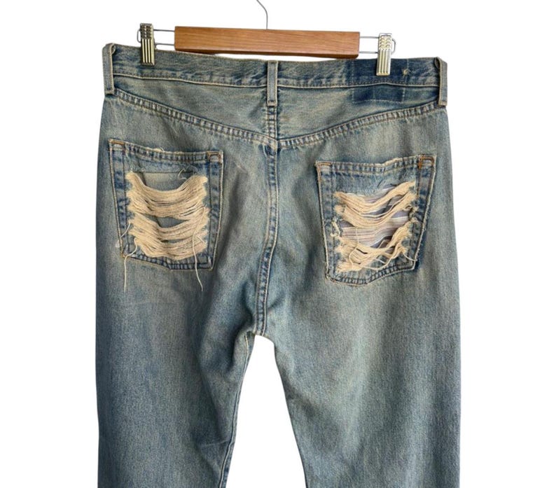 May include: Light blue denim jeans with distressed detailing on the back pockets. The jeans are hanging on a wooden hanger. The pockets have a shredded design, revealing the fabric underneath. The jeans are a classic, casual style.