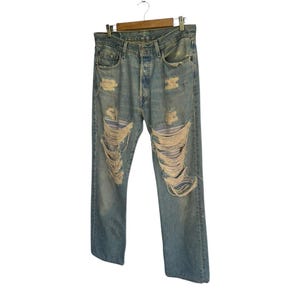 May include: Light blue denim jeans with distressed detailing, including large rips and frayed edges. The jeans are hanging on a wooden hanger against a white background. The jeans have a classic five-pocket design.