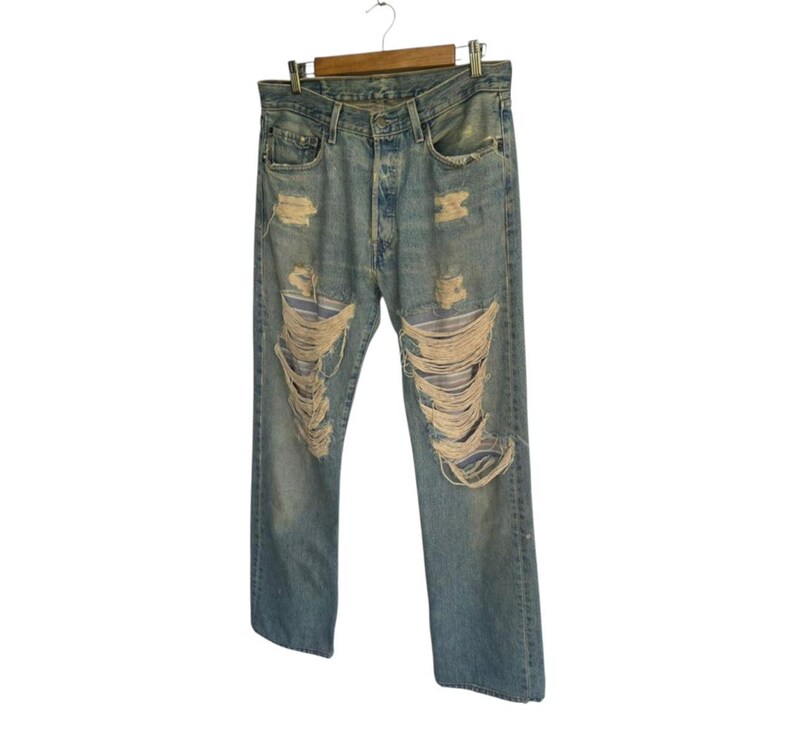 May include: Light blue denim jeans with distressed detailing, including large rips and frayed edges. The jeans are hanging on a wooden hanger against a white background. The jeans have a classic five-pocket design.