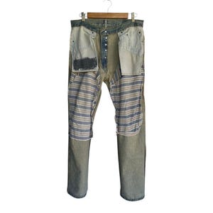 May include: Distressed denim jeans with a unique design. The jeans feature a light wash with visible wear and tear. Patches of striped fabric in blue, gray, and white are sewn into the front. The jeans are displayed on a wooden hanger.