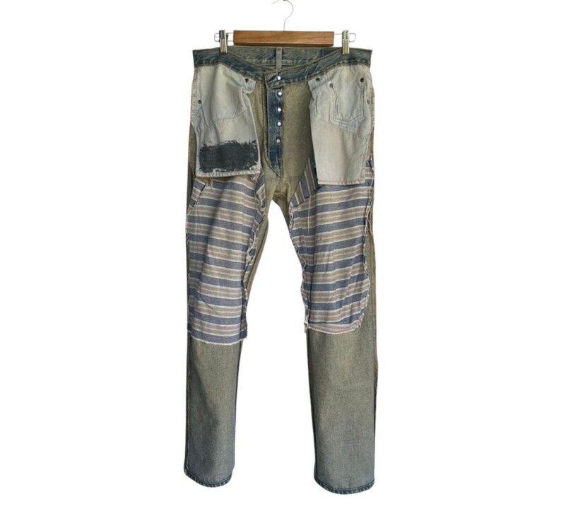 May include: Distressed denim jeans with a unique design. The jeans feature a light wash with visible wear and tear. Patches of striped fabric in blue, gray, and white are sewn into the front. The jeans are displayed on a wooden hanger.