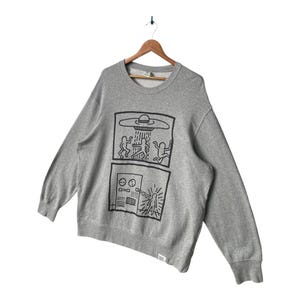 May include: A gray crewneck sweatshirt with a black graphic design featuring two panels. The top panel depicts a UFO with figures, and the bottom panel shows a control panel with a beam. The sweatshirt is hanging on a wooden hanger.