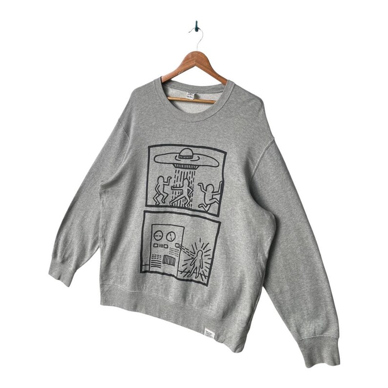 May include: A gray crewneck sweatshirt with a black graphic design featuring two panels. The top panel depicts a UFO with figures, and the bottom panel shows a control panel with a beam. The sweatshirt is hanging on a wooden hanger.