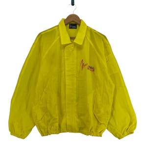 May include: A bright yellow vintage jacket with a collared neckline, two front pockets, and elasticated cuffs and hem. The jacket features a red logo on the chest that reads "New Era". It is hanging on a wooden hanger.