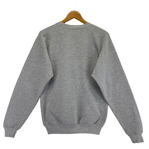 May include: A grey sweatshirt with a crew neck and long sleeves, suspended from a wooden hanger. The rear view of the garment reveals its plain, classic design. The material looks soft and comfortable. The colour is a medium grey.