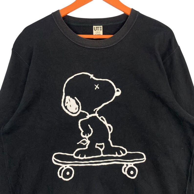 May include: Black crew neck sweatshirt featuring a white graphic of Snoopy skateboarding. The design includes a white outline of Snoopy with an "X" for an eye and a skateboard. The sweatshirt has a ribbed neckline.