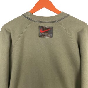 May include: Olive green Nike sweatshirt with a crew neck and long sleeves. Features a rectangular patch on the upper back with a red Nike swoosh logo and the text "X-TRAINING". The sweatshirt is hanging on an orange hanger.