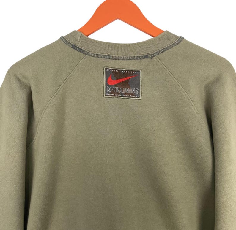 May include: Olive green Nike sweatshirt with a crew neck and long sleeves. Features a rectangular patch on the upper back with a red Nike swoosh logo and the text "X-TRAINING". The sweatshirt is hanging on an orange hanger.