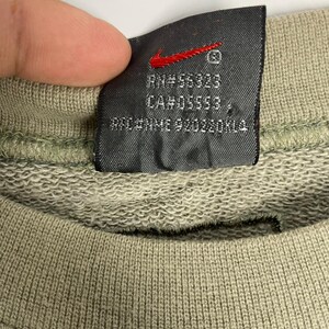 May include: Close-up of a tag from a Nike sweatshirt. The tag is dark gray with a red Nike swoosh logo. Text on the tag includes RN#56323, CA#05553, and RFC/NMME 920220KLA. The sweatshirt is olive green.