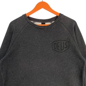 Pode incluir: Um moletom cinza escuro com a palavra "DEUS" em um design geom&eacute;trico preto na frente. O moletom tem gola redonda e mangas compridas. A pe&ccedil;a de roupa est&aacute; pendurada em um cabide laranja.