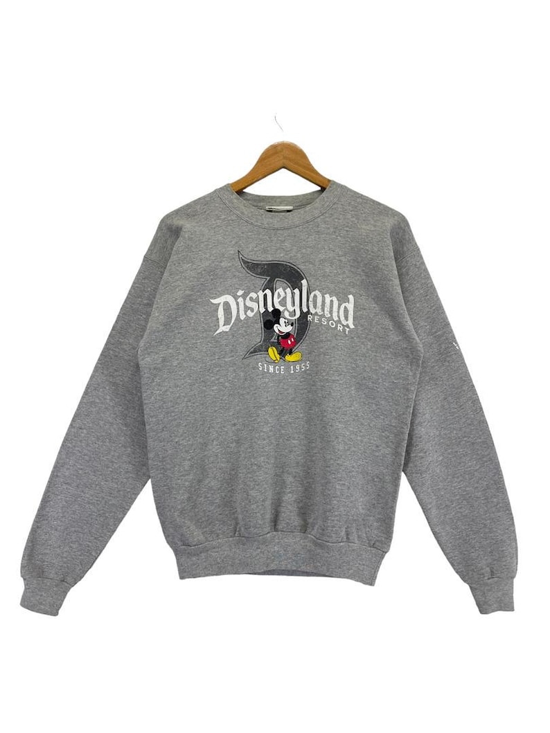 May include: A grey sweatshirt featuring the Disneyland Resort logo and a Mickey Mouse graphic. The logo is in a stylized font with a large letter "D". The sweatshirt has long sleeves and a crew neck.