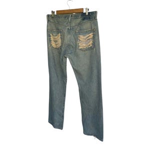 May include: Light blue denim jeans with distressed detailing on the back pockets. The jeans are hanging on a wooden hanger against a white background. The jeans have a classic five-pocket design.