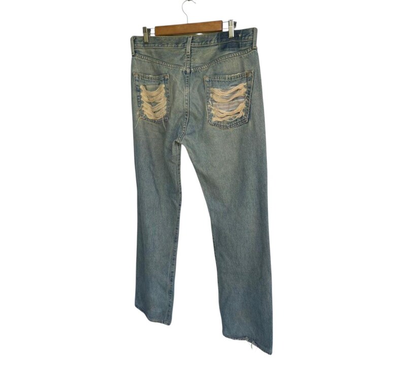 May include: Light blue denim jeans with distressed detailing on the back pockets. The jeans are hanging on a wooden hanger against a white background. The jeans have a classic five-pocket design.