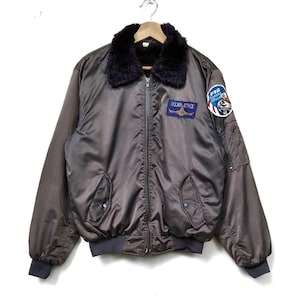 May include: A brown bomber jacket with a black faux fur collar. The jacket features a zip closure, two front pockets, and patches that read "US AIR FORCE" and "F16 Fighting Falcon". It has dark grey ribbed cuffs and waistband.