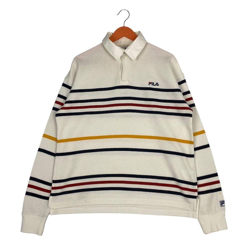 May include: A long-sleeve polo shirt in off-white featuring horizontal stripes in navy blue, dark red, and mustard yellow. The shirt has a collared neckline with a button placket and the FILA logo embroidered in navy blue on the upper left chest.