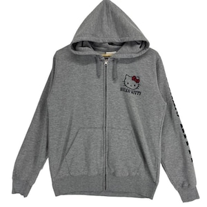 May include: A gray zip-up hoodie featuring the Hello Kitty character with a red bow. The hoodie has a hood, a front pocket, and the words "HELLO KITTY" printed below the character. The sleeve has a black graphic design.