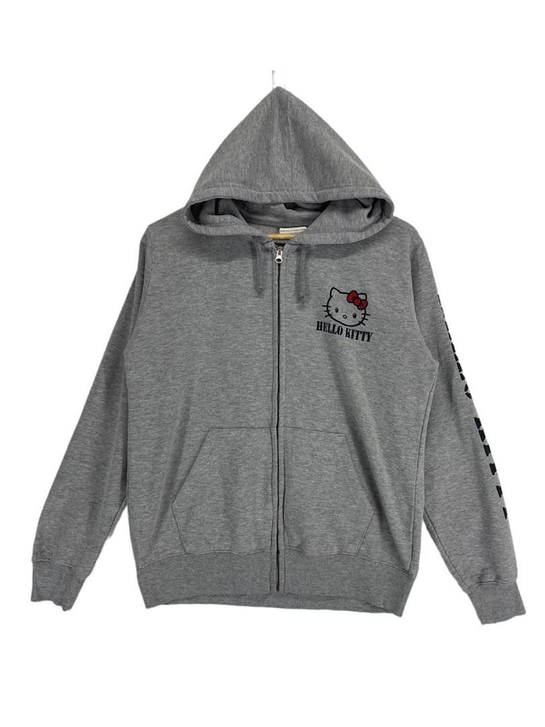 May include: A gray zip-up hoodie featuring the Hello Kitty character with a red bow. The hoodie has a hood, a front pocket, and the words "HELLO KITTY" printed below the character. The sleeve has a black graphic design.