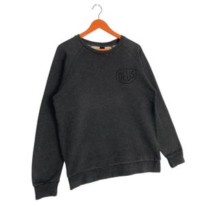 Pode incluir: Camisola cinza escuro com a palavra "DEUS" em fonte preta e estilizada no peito. A camisa de manga comprida e gola redonda est&aacute; pendurada em um cabide de madeira laranja. A camisola &eacute; feita de um material macio.