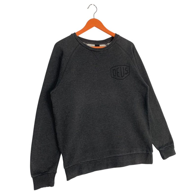 Pode incluir: Camisola cinza escuro com a palavra "DEUS" em fonte preta e estilizada no peito. A camisa de manga comprida e gola redonda est&aacute; pendurada em um cabide de madeira laranja. A camisola &eacute; feita de um material macio.