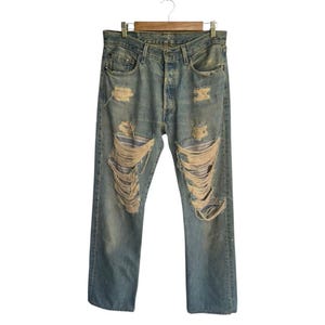 May include: Light blue denim jeans with distressed detailing, including large rips across the front of the legs. The jeans are hanging on a wooden hanger, showcasing a classic, worn aesthetic.