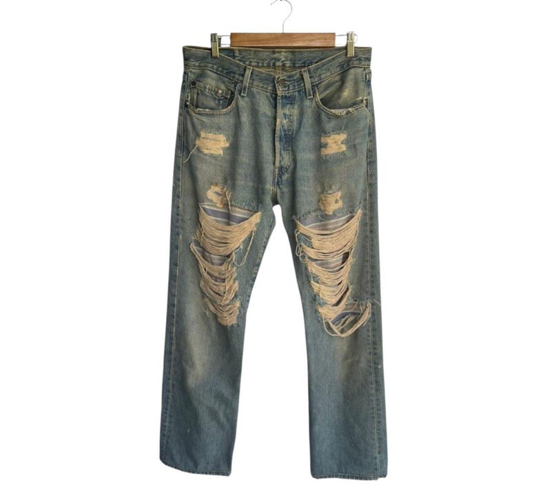 May include: Light blue denim jeans with distressed detailing, including large rips across the front of the legs. The jeans are hanging on a wooden hanger, showcasing a classic, worn aesthetic.