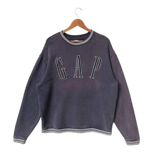 Peut inclure: Un sweat-shirt GAP bleu marine vintage avec le nom de la marque en lettres blanches. Le sweat-shirt a un col rond et des poignets et un ourlet côtelés avec des rayures blanches et noires. Le vêtement est suspendu à un cintre en bois.