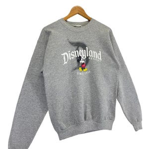May include: A grey Disneyland Resort sweatshirt with long sleeves. The front displays the Disneyland logo in white and black, with Mickey Mouse and the text "SINCE 1955". The sweatshirt is suspended on a wooden hanger.