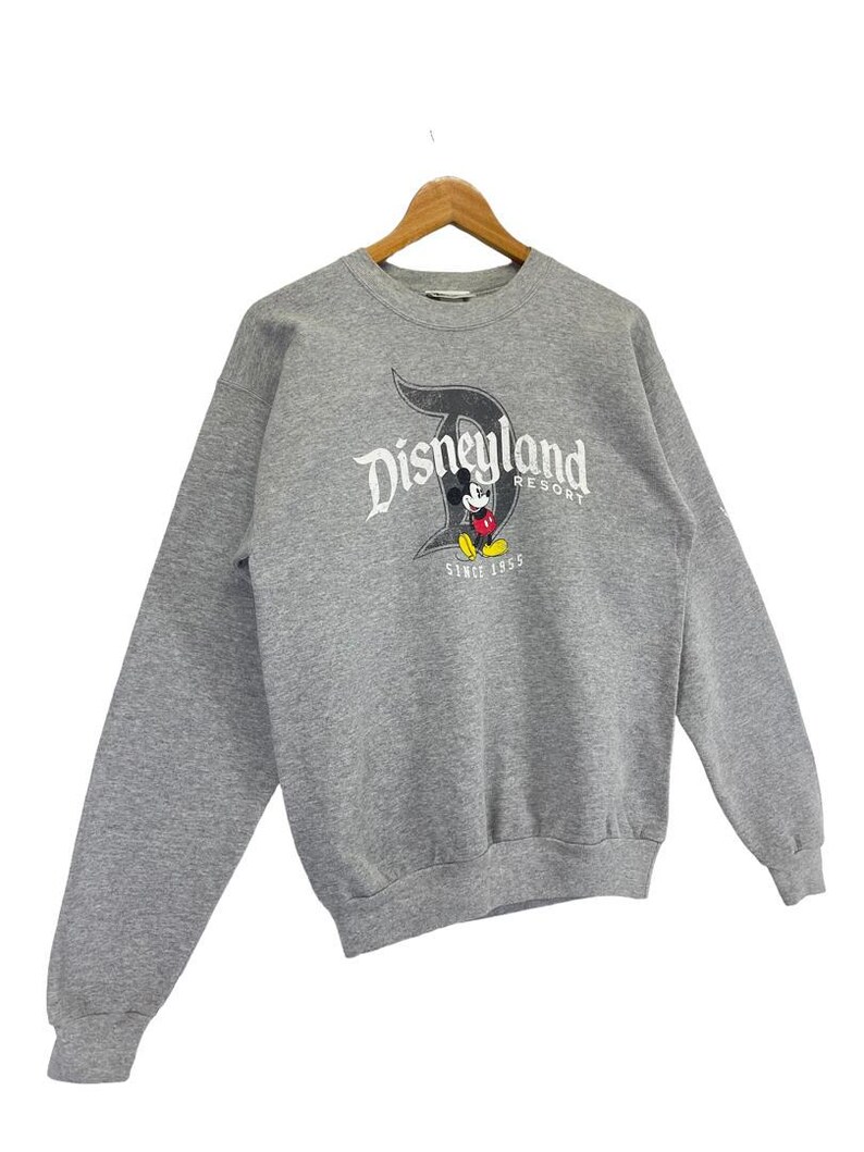 May include: A grey Disneyland Resort sweatshirt with long sleeves. The front displays the Disneyland logo in white and black, with Mickey Mouse and the text "SINCE 1955". The sweatshirt is suspended on a wooden hanger.