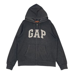 Könnte beinhalten: Dunkelgrauer Kapuzenpullover mit Reißverschluss, Kapuze und Vordertasche. Das Wort "GAP" ist in weißen Blockbuchstaben auf der Brust abgebildet. Der Hoodie hat einen durchgehenden Reißverschluss und lange Ärmel.
