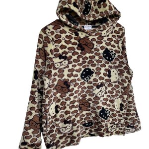 Może przedstawiać: Brązowa i beżowa bluza z kapturem z polaru z wzorem w cętki i postaciami Hello Kitty. Bluza z kapturem ma kaptur i długie rękawy. Postacie Hello Kitty są w kolorze czarnym, brązowym i białym.