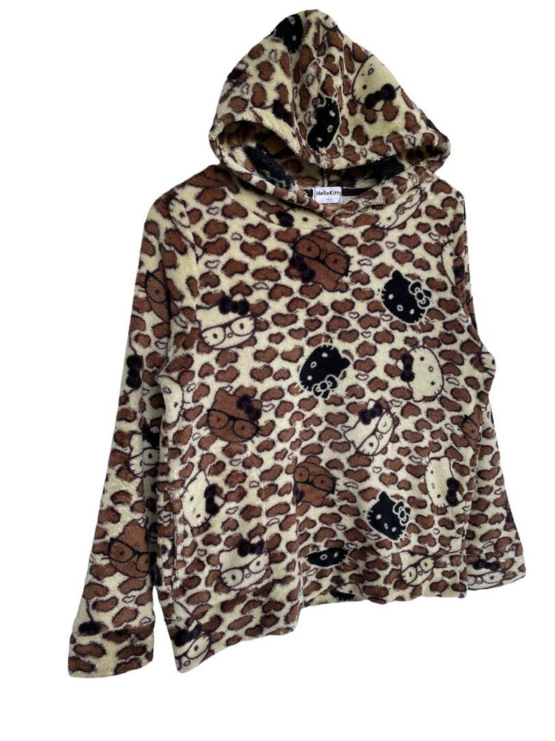 Może przedstawiać: Brązowa i beżowa bluza z kapturem z polaru z wzorem w cętki i postaciami Hello Kitty. Bluza z kapturem ma kaptur i długie rękawy. Postacie Hello Kitty są w kolorze czarnym, brązowym i białym.