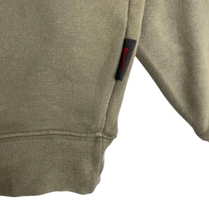 May include: Close-up of a sage green sweatshirt. The garment features a ribbed hem and cuff. A small black tag with a red Nike swoosh logo is sewn on the sleeve. The fabric appears to be a soft, comfortable material.