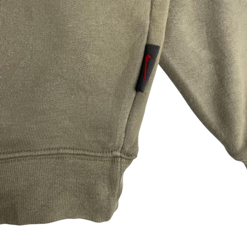 May include: Close-up of a sage green sweatshirt. The garment features a ribbed hem and cuff. A small black tag with a red Nike swoosh logo is sewn on the sleeve. The fabric appears to be a soft, comfortable material.