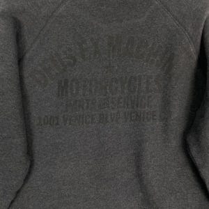 Pode incluir: Camisola cinza escuro com o texto "DEUS EX MACHINA MOTORCYCLES PARTS & SERVICE 1001 VENICE BLVD VENICE CA" impresso em cinza mais escuro. A camisola tem gola redonda e um aspeto macio e texturizado.