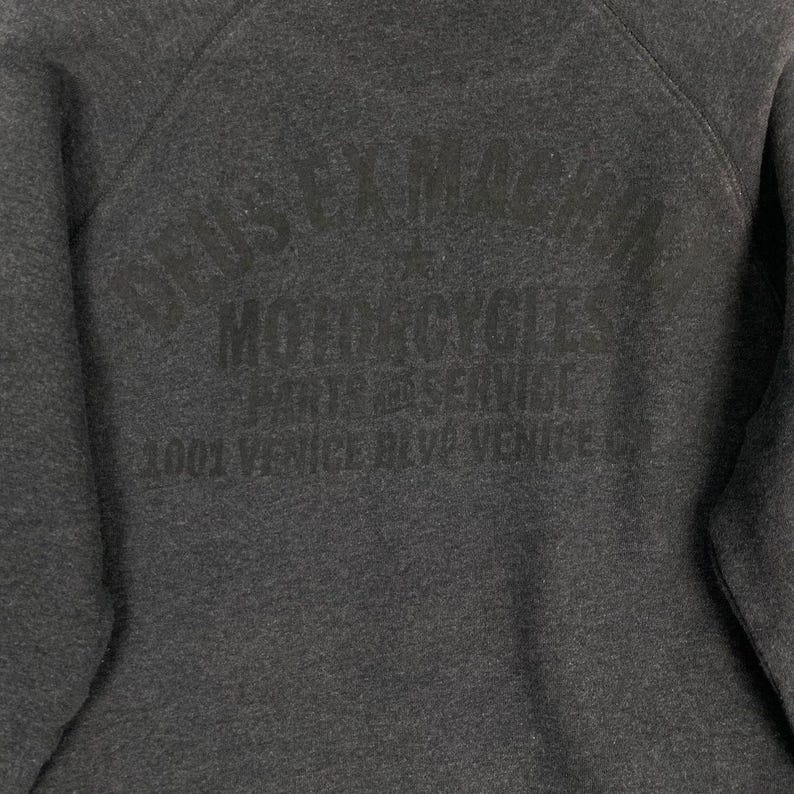 Pode incluir: Camisola cinza escuro com o texto "DEUS EX MACHINA MOTORCYCLES PARTS & SERVICE 1001 VENICE BLVD VENICE CA" impresso em cinza mais escuro. A camisola tem gola redonda e um aspeto macio e texturizado.